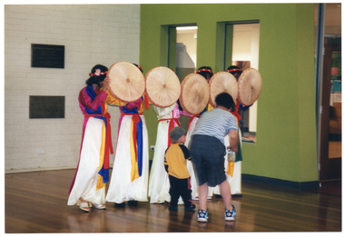 COL photo of a man and a child standing close to a troupe of dancers in traditional Vietnamese dress. The man is bending forward as he holds the child's hand. The dancers are each holding a round, flat-bottomed basket in front of their faces.
