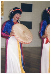 COL close up photo of a dancer in traditional dress (with another dancer partially obscured). She is holding a round, flat-bottomed basket.