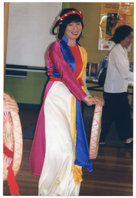 COL close up photo of a dancer in traditional dress (with another dancer partially obscured). She is holding a round, flat-bottomed basket. In the background, there is another woman (in contemporary casual dress) walking past.