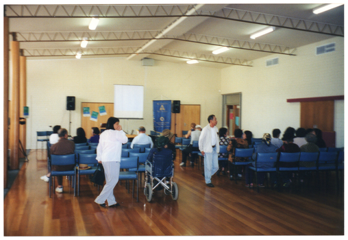 COL photo of a small group of people gathered inside a hall. Most people are seated on rows of chairs. One person is a wheelchair user. Two people are walking around. In the background, there are two speakers on tall vertical stands, with a projector screen and a large blue and gold banner on a tall vertical stand between them. There are two sets of double wooden doors on either side of the projector screen and another glass door on a perpendicular wall.