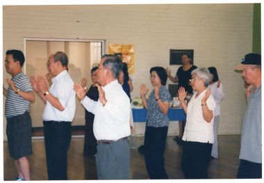 COL photo of a group of people taking part in an exercise session in a large room. They are holding their hands up, palms outwards. In the background, there is a woman standing behind a table display with food products. She is also taking part in the exercise session. There are some posters on the wall behind her.