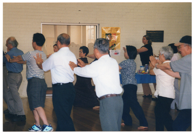 COL photo of a group of people taking part in an exercise session in a large room. They have their arms outstretched, palms outwards, as they twist their bodies to one side. In the background, there is a woman standing behind a table display with food products. She is also taking part in the exercise session. There are some posters on the wall behind her.