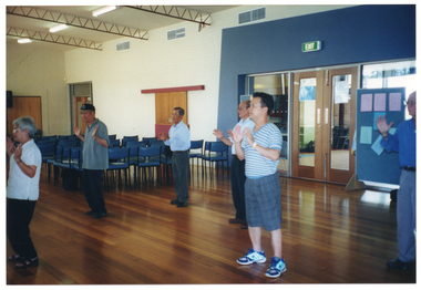 COL photo of a group of people taking part in an exercise session in a large room. They are holding their hands up, palms outwards. From the centre left of the frame, there are several rows of empty chairs. Behind them, there are two freestanding vertical display boards and a set of closed double glass doors. There is another glass door towards the back of the hall.