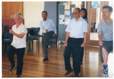 COL photo of a group of people taking part in an exercise session in a large room. They are standing on one leg as they lift the other leg, bending at the knee. To the left of the frame, there are several rows of empty chairs. Behind them, there is a freestanding vertical display board and a set of closed double glass doors.