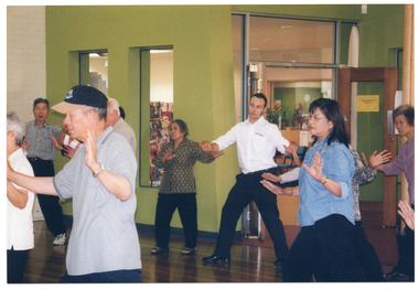 COL photo of a group of people taking part in an exercise session in a large room. They are standing with their feet wide apart and hands up, palms facing outwards. In the background, there are some glass panels in the wall. There is also another room accessible through a wide opening in the wall.