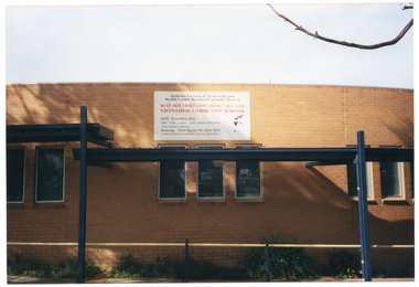 COL photo of a large sign affixed high up on the exterior brick wall of a building. It reads "Arthritis Victoria & Western Region Health Centre Braybrook proudly present BUÔ'I HÔI THA'O CÔNG DÔNG VIÊT NAM VIETNAMESE COMMUNITY SEMINAR DATE: 18 MARCH 2003 TIME: 10.00 - 3.00pm COST: GOLD COIN ENTRY Guest speakers attending Bookings - Trinh Nguyen Ph: 9364 9622 A light luncheon is available (free) for participants". There are also three logos on the bottom right side of the sign, one of which is the Arthritis Victoria logo.