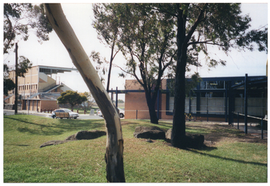 COL wide angle photo of the exterior of two large buildings, one of which appears to be part of a grandstand. There is a small car park between the two buildings, with two cars parked there.