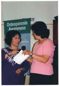 COL photo of two women standing in front of a freestanding green and blue Osteoporosis Awareness banner. One of them is holding a microphone.