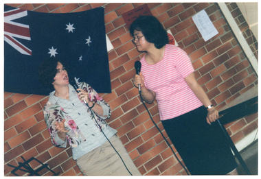 COL photo of two women, standing. They are each holding a hand-held a microphone. Behind them is an Australian flag hanging on a brick wall.