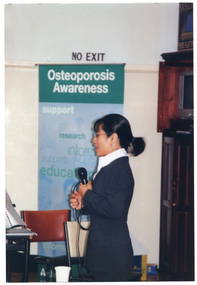 COL photo of a woman, standing, speaking into a microphone. Beside her is a freestanding green and blue Osteoporosis Awareness banner.
