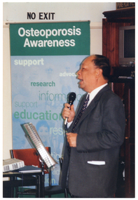 COL photo of a man, standing, speaking into a microphone. Beside him is a freestanding green and blue Osteoporosis Awareness banner.