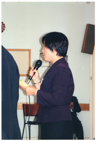 COL photo of a woman, standing, speaking into a microphone. One other person (partially obscured) is standing nearby.