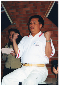 COL photo of a man demonstrating some arm exercises. He is sitting on the edge of a table. Behind him is a woman speaking into a microphone. On the brick wall are two Australian flags.