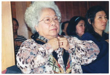 COL close up photo of an elderly woman taking part in an exercise session, with other participants in the background. Everyone is standing up and pushing their fists together in front of their chests. In the background, there are several wooden honour boards up high on the wall.