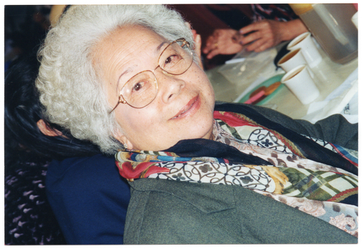 COL close up photo of an elderly woman sitting at a table. On the table, there are some disposable cups of coffee. A few other people (partially obscurred) are also sitting at the table. The photo has been taken at a 45 degree angle.