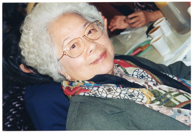 COL close up photo of an elderly woman sitting at a table. On the table, there are some disposable cups of coffee. A few other people (partially obscurred) are also sitting at the table. The photo has been taken at a 45 degree angle.