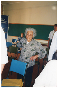 COL photo of an elderly woman taking part in a group exercise inside a hall. She is standing behind a chair, with her hands on her hips. Other people (partially obscured) are also taking part in the exercise session.