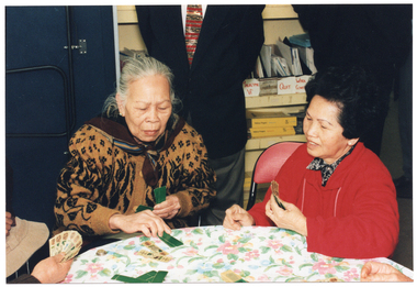 COL photo of two women sitting at a round table covered with a floral tablecloth. They are playing a traditional playing card game with two other people (partially obscured), also seated at the table. The cards are long and narrow. Two other people, standing behind them (partially obscured), look on. In the background, there is some shelving containing several magazine holders filed with paperwork and several Yellow Pages telephone directories.