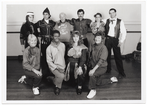 B&W photo of a group of people posing for a Photograph.  Four people in the back row are standing, while four people in the front row are kneeling on one knee. Several people are dressed in traditional costumes, with one young man wearing a plain tunic top and slacks with a kerchief around his neck. Three women are wearing sweatshirts with the text "Move It Or Lose It" and a logo printed on the front.