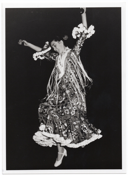 B&W photo of a flamenco dancer, in traditional dress, dancing while playing the castanets. She has both arms held up high above her head while one foot is off the ground.
