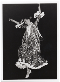 B&W photo of a flamenco dancer, in traditional dress, dancing while playing the castanets. She has both arms held up high above her head while one foot is off the ground.