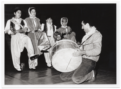 B&W photo of four dancers, dressed in traditional costumes, standing on one foot while lifting the other foot. Kneeling in front of them, a musician (dressed in casual clothes) plays a large drum.