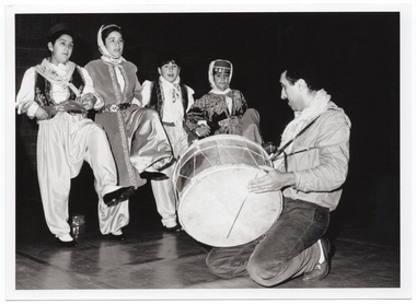 B&W photo of four dancers, dressed in traditional costumes, standing on one foot while lifting the other foot. Kneeling in front of them, a musician (dressed in casual clothes) plays a large drum.