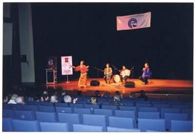 COL photo of a four-piece music and dance troupe performing on a large stage. The troupe consists of a female dancer and three musicians: a dambooka/darabukka drummer, a piano accordion player, and a violinist . They are all wearing Middle-Eastern inspired traditional clothing/costumes. The dancer is holding her arms out wide, mid-dance, while the musicians are seated. Above the stage, affixed to a large black backdrop, is a large white banner with a blue logo and the text, "Ethnic Communities' Council of Victoria". Beside the troupe is a freestanding vertical banner on a stand, with the text, "ACTIVE FOR LIFE JUST FIND 30 MINS A DAY", and a corresponding logo. Beside it is another banner with a logo and text in white on a dark blue background. The text heading is "Victoria 1999". There is a small audience watching the troupe from the auditorium seating.