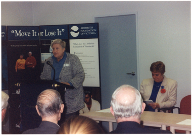 COL photo of a woman standing at a black lectern, speaking into a microphone. Beside her, there is another woman sitting at a long rectangular table. Behind her, there is a freestanding display board with the heading, "Move It or Lose It", the Arthritis Foundation of Victoria text and logo, and various posters and information. In the foreground, there are a few people sitting in the audience (partially obscured), facing them.