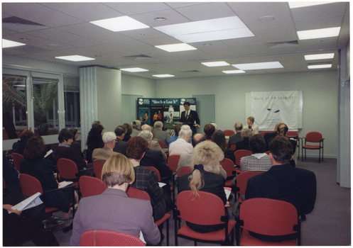 COL photo of a man, standing at a lectern, speaking to a large group of people (seated) in a large room. Beside him, there is a woman seated at a table, facing the group. Behind them is a large freestanding Arthritis Foundation of Victoria display board and a large white and blue banner draped across a whiteboard fixed to the wall. Both the display board and the banner contain the text "Move It or Lose It" as a heading. The room contains bi-fold doors, which have been folded back to connect two rooms into one. On the left of the frame, there are some large windows through which a small palm tree is visible.
