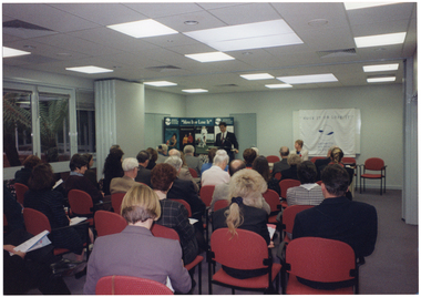 COL photo of a man, standing at a lectern, speaking to a large group of people (seated) in a large room. Beside him, there is a woman seated at a table, facing the group. Behind them is a large freestanding Arthritis Foundation of Victoria display board and a large white and blue banner draped across a whiteboard fixed to the wall. Both the display board and the banner contain the text "Move It or Lose It" as a heading. The room contains bi-fold doors, which have been folded back to connect two rooms into one. On the left of the frame, there are some large windows through which a small palm tree is visible.