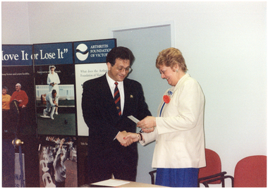 COL photo of a man and a woman, (both standing), shaking hands. The woman is holding a small piece of paper and they are both smiling. Behind them is an Arthritis Foundation of Victoria "Move It Or Lose It" display board with information and posters.