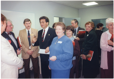 COL photo of a group of people standing in a room. Many of them are holding one or more large printed programs with a red cover.