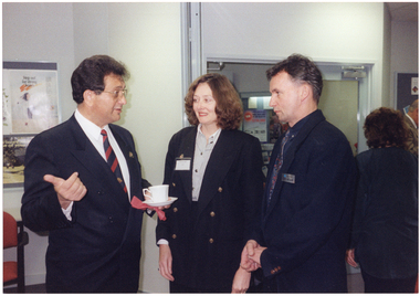 COL photo of three people standing close to one another, chatting. One of them is holding a white cup and saucer in one hand while gesticulating with the other. In the background, there are a few people standing to the right of the frame.