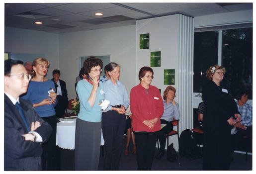 COL photo of a group of people gathered in a large room. Many of them are standing and a few are sitting on chairs. Some people are holding a glass in one hand. In the background, there is a table covered with a white tablecloth. A set of bi-fold doors are folded against the wall to connect two rooms together. There are three green posters on the outermost door panel. There are some large windows along the wall, through which it is evident that it is dark outside.