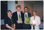 COL photo of four people standing close together, in a row. Three of them are each holding a plastic video case with a green cover while the other person is holding a large folder, also with a green cover. The covers of the folder and video cases match the posters on the open bi-fold door panel behind them. There is a large window beside the open bi-fold doors.