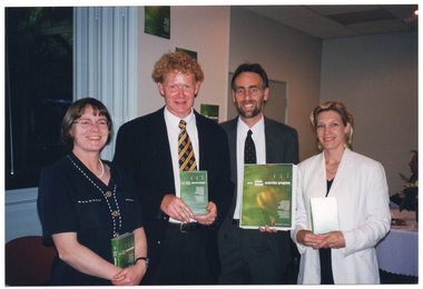 COL photo of four people standing close together, in a row. Three of them are each holding a plastic video case with a green cover while the other person is holding a large folder, also with a green cover. The covers of the folder and video cases match the posters on the open bi-fold door panel behind them. There is a large window beside the open bi-fold doors.