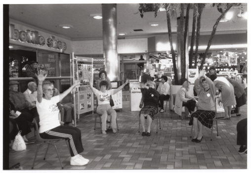 B&W photo of a group of people sitting on some chairs at a shopping centre. The chairs have been set up in a semi-circle in an open area in front of some shops. Behind them, there is a large freestanding Arthritis Foundation of Victoria display board and several palm trees in a large pot. The display board contains information and several Photographs. In the background, there are some shops and shoppers visible. Above one shop is a sign, consisting of individual letters that are backlit, which spells out the words, "bottle shop".