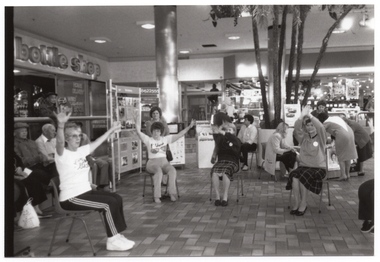 B&W photo of a group of people sitting on some chairs at a shopping centre. The chairs have been set up in a semi-circle in an open area in front of some shops. Behind them, there is a large freestanding Arthritis Foundation of Victoria display board and several palm trees in a large pot. The display board contains information and several Photographs. In the background, there are some shops and shoppers visible. Above one shop is a sign, consisting of individual letters that are backlit, which spells out the words, "bottle shop".