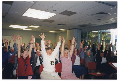 COL photo of a large group of people seated on rows of chairs in a large room. They all have their arms outstretched above their heads. In the background, a set of bi-fold doors, which have been folded up in the open position, are visible beside some large windows. Various shrubs are visible through the windows.
