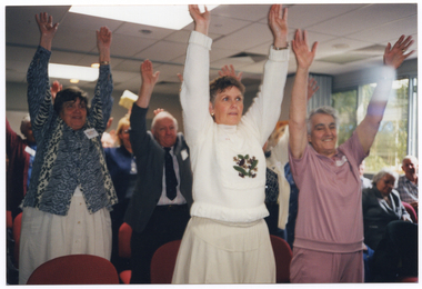 COL photo of a large group of people in a large room. Several people are standing in front of a chair, which have been set up in rows, while others remain seated. The people standing all have their arms outstretched above their heads. In the background, a set of bi-fold doors, which have been folded up in the open position, are visible beside some large windows. Various shrubs are visible through the windows.
