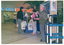 COL photo of several people standing around an information stall at a shopping centre. There are a few people standing behind the stall table and two people perusing information. One man is standing near the stall in front of a freestanding display board and a small colour television on a cart. He is holding a leaflet. Affixed to the display board is a National Arthritis Week poster featuring television presenter, Tony Barber, amongst Photographs and information. The same poster appears affixed on the side of the tablecloth covering the stall table. In the background, there is a supermarket with a partially obscured sign "...EW WORLD".