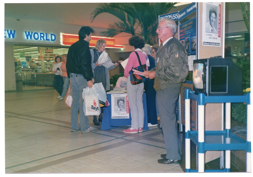 COL photo of several people standing around an information stall at a shopping centre. There are a few people standing behind the stall table and two people perusing information. One man is standing near the stall in front of a freestanding display board and a small colour television on a cart. He is holding a leaflet. Affixed to the display board is a National Arthritis Week poster featuring television presenter, Tony Barber, amongst Photographs and information. The same poster appears affixed on the side of the tablecloth covering the stall table. In the background, there is a supermarket with a partially obscured sign "...EW WORLD".