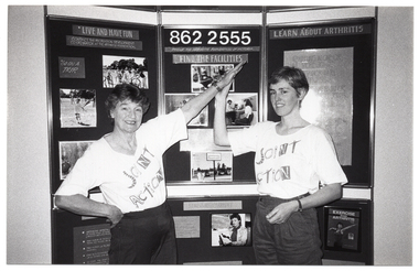 B&W photo of two women stand in front of a freestanding multi-panel Arthritis Foundation of Victoria display board. They are wearing t-shirts with a graphic design that spells out the words “Joint Action”.