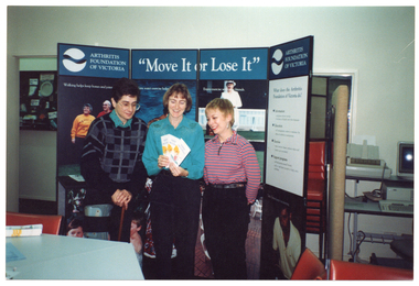 COL photo of three people standing in front of a large freestanding display board. One of them is holding some information leaflets in her hand. To one side of the display board is a desk or trolley with some computer equipment and a printer. There is a stack of chairs against the wall, behind the display board. In the foreground, there are two tables, joined together, and a chair.