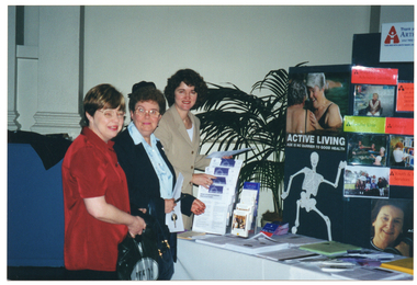 COL photo of three women standing next to an information stall and display board. They are looking at the camera and smiling. On the table, there are various information leaflets and factsheets, some of which are displayed on clear perspex brochure stands. Behind the table, there is a large black display board containing posters and large Photographs. One of the posters, featuring a drawing of a skeleton, is titled "Active Living", with the subtitle, "age is no barrier to good health". Other images featured on the display board include photos relating to various programs offered by the AFV, including the Telephone Advisory Service, water exercise classes (under the title 'Exercise Unit'), the Library, and Youth & Family Services. At the top of the display board, there is an Arthritis Victoria sign. Beside the table and display board, there is a large potted palm plant.