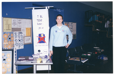 COL photo of a woman standing next to a small display table. Behind the table, there is a freestanding vertical Vic Health and Arthritis Victoria banner. The banner is titled 'TABLE TENNIS keeps you ACTIVE FOR LIFE' and features the Active for Life, Vic Health and Arthritis Victoria logos. On the wall behind her, there is a display board on either side featuring various posters relating to table tennis. Next to the banner are some chairs, on which there is a stack of posters and folders.