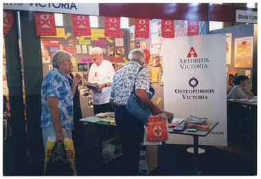 COL photo of two people perusing an information stall at an expo event. There are two people (one partially obscured) standing behind the stall tables. At the front of the stall is a large freestanding 'Arthritis Victoria and Osteoporosis Victoria' banner, featuring their respective logos. There is some red bunting strung across the top of the front of the stall, containing the VicHealth 'Active For Life' text and logo. At the back of the stall, to the left, is a poster featuring media personality, Ita Buttrose: it is titled 'Invest in your Future'.