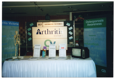 COL photo of an Arthritis Victoria information stall at an unidentified event. The stall consists of a display board and table in the centre, flanked by two large blue and green freestanding banners on either side. One banner is titled, "Arthritis Victoria", while the other is titled, "Osteoporosis Awareness". Behind the table, thre is a display board featuring the stall number '103' and a sign that says 'ARTHRITIS VICTORIA'. Beneath that are two large signs - "Arthritis Victoria" and "Osteoporosis Victoria", with their respective logos. On either side of the large signs are some small printed cards affixed to display board behind the stall. The stall table is covered with a white, floor-length tablecloth. On the table, there are various brochures and booklets, a small skeleton on a stand, and a small portable television set (which is displaying some information).