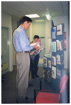 COL photo of three people (one obscured) standing in front of a book display. The books are contained inside clear perspex holders, which have been affixed to a freestanding display board. Each person is looking down at an open book, which they are holding in their hands.